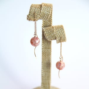 May include: A pair of drop earrings featuring spherical pink beads with a pearlescent, mosaic-like finish. Each bead is suspended from a thin gold-toned chain attached to a curved gold-toned wire hook, creating a delicate and dangling silhouette.