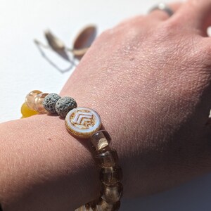 May include: A beaded bracelet featuring a mix of amber, yellow, and gray beads. The bracelet includes square and round beads, with a central white and gold circular bead. The bracelet is worn on a wrist.