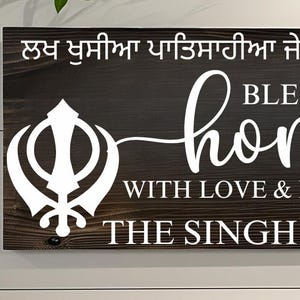 May include: A rectangular wooden sign with the text "BLESS OUR home WITH LOVE & LAUGHTER THE SINGH FAMILY" in white lettering. The sign also features two white Sikh symbols. The background is a dark brown.