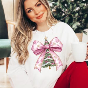 May include: White sweatshirt with a watercolour Christmas tree design. The tree is decorated with pink baubles and a star, set within a large pink bow. The design includes gold accents and snowflake details, ideal for the festive season.