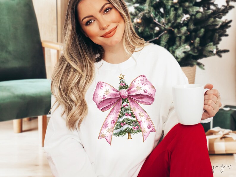 May include: White sweatshirt with a watercolour Christmas tree design. The tree is decorated with pink baubles and a star, set within a large pink bow. The design includes gold accents and snowflake details, ideal for the festive season.