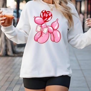 May include: White sweatshirt with a pink balloon animal design. The balloon animal is shaped like a poodle and has a red heart-shaped balloon on its head. The poodle is wearing a pink bow.