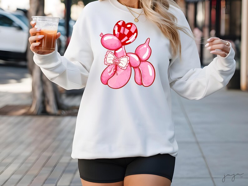 May include: White sweatshirt with a pink balloon animal design. The balloon animal is shaped like a poodle and has a red heart-shaped balloon on its head. The poodle is wearing a pink bow.
