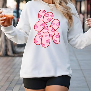May include: White sweatshirt with a pink and red graphic of a balloon animal shaped like a dog with hearts on it.