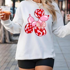 May include: White sweatshirt with a red and pink checkered balloon dog graphic. The balloon dog has a pink bow.