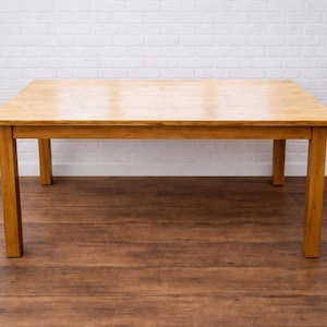 May include: A rectangular wooden table with four legs. The table is made of light-coloured wood and has a smooth, polished surface. The table is set against a white brick wall and a wooden floor.
