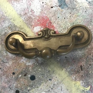 May include: A gold-toned metal drawer pull with a decorative design. The pull has two loops on either side and a raised centre section.