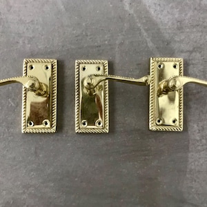 May include: Three brass door handles with rectangular backplates and rope-style edging. The handles feature a curved lever design. The hardware is a shiny gold colour.
