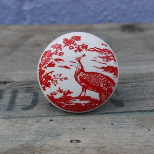 May include: White ceramic knob featuring a red peacock design. The peacock is standing on a branch with red flowers.