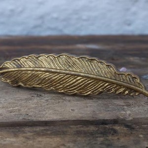 May include: A gold-coloured metal feather-shaped drawer pull. The pull has a detailed design and is perfect for adding a touch of elegance to any home decor.