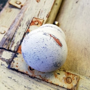 May include: A white painted wooden knob with a distressed finish, revealing some of the original wood beneath. The knob is attached to a piece of weathered wood with rusty metal fittings.