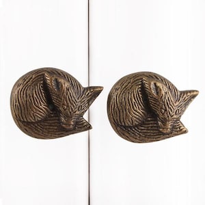 May include: Two antique gold-toned metal cabinet knobs shaped like sleeping foxes. The foxes are curled up with their tails wrapped around their bodies.
