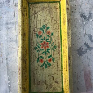 May include: A rectangular wooden tray with a distressed yellow paint finish. The tray has a green border and a floral design painted in the centre with orange, red, and green accents.