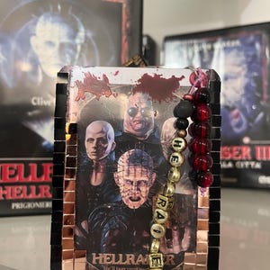Może przedstawiać: Obraz Hellraiser w mozaikowej ramie z koralikowym urokiem. Grafika przedstawia postacie z horroru z tytułem "Hellraiser" i hasłem "He'll tear your soul apart". Urok wymawia "HELLRAISER".