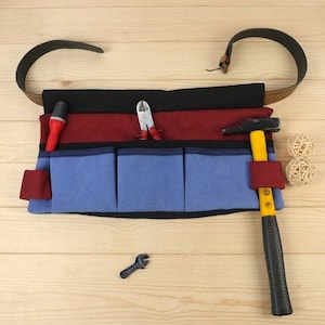 Peut inclure: Un tablier à outils multi-poches avec une ceinture noire, une section centrale bordeaux et trois poches avant bleu clair. Le tablier contient un outil à manche rouge, une pince en métal et un marteau à manche jaune, avec une petite clé en dessous.