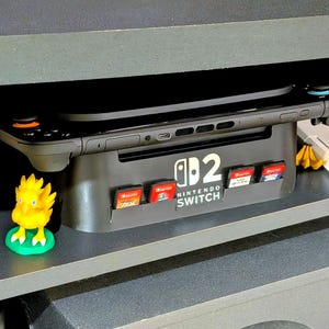 May include: A black Nintendo Switch console rests on a gray stand with game cartridge slots. The stand has the Nintendo Switch logo. Several game cartridges are inserted. A yellow Chocobo figurine sits on the shelf below. The console is on a shelf with a white box with red accents.