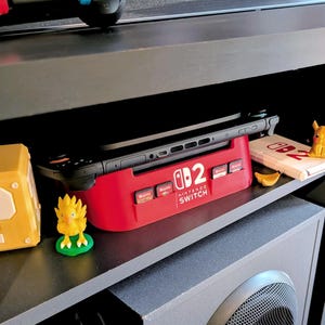 Może przedstawiać: Czerwona stacja dokująca Nintendo Switch z napisem "Nintendo Switch". Mieści czarną konsolę Nintendo Switch. Widoczny jest r&oacute;wnież ż&oacute;łty blok pytajnika Super Mario i figurka Pikachu.