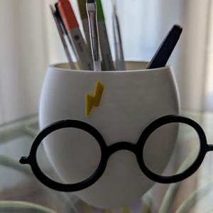 May include: White ceramic pencil holder shaped like Harry Potter's face with black round glasses and a yellow lightning bolt on the forehead.