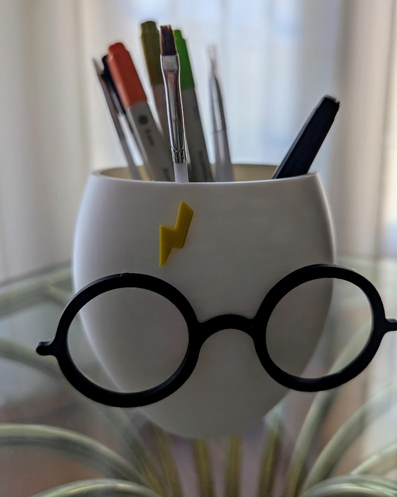 May include: White ceramic pencil holder shaped like Harry Potter's face with black round glasses and a yellow lightning bolt on the forehead.