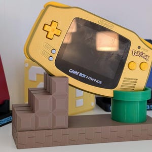 May include: A yellow Game Boy Advance console with "Pokémon" and "Game Boy Advance" text, resting on a brown platform with a green pipe and brown steps. Other consoles are visible in the background.