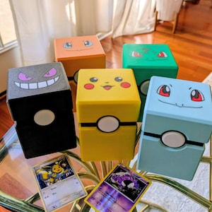 Könnte beinhalten: Eine Sammlung farbenfroher, würfelförmiger Aufbewahrungsboxen mit Designs beliebter Zeichentrickfiguren. Die Boxen sind in verschiedenen Farben gehalten, darunter Gelb, Schwarz, Orange, Grün und Blau. Zwei Sammelkarten sind im Vordergrund zu sehen.