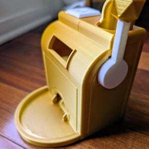 Könnte beinhalten: Ein goldfarbener 3D-gedruckter Würfelturm mit einem weißen Griff und einem Tablett am Boden. Der Turm hat einen Schlitz zum Würfeln und eine durchsichtige Öffnung, um die Würfel zu sehen.