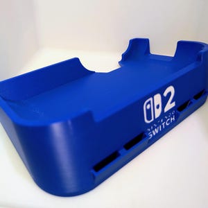 Może przedstawiać: Żywy niebieski dock Nintendo Switch. Dock posiada logo Nintendo Switch i cyfrę 2 w kolorze białym. Projekt obejmuje tacę i dwa podwyższone podpory. Widoczne jest r&oacute;wnież słowo "SWITCH".
