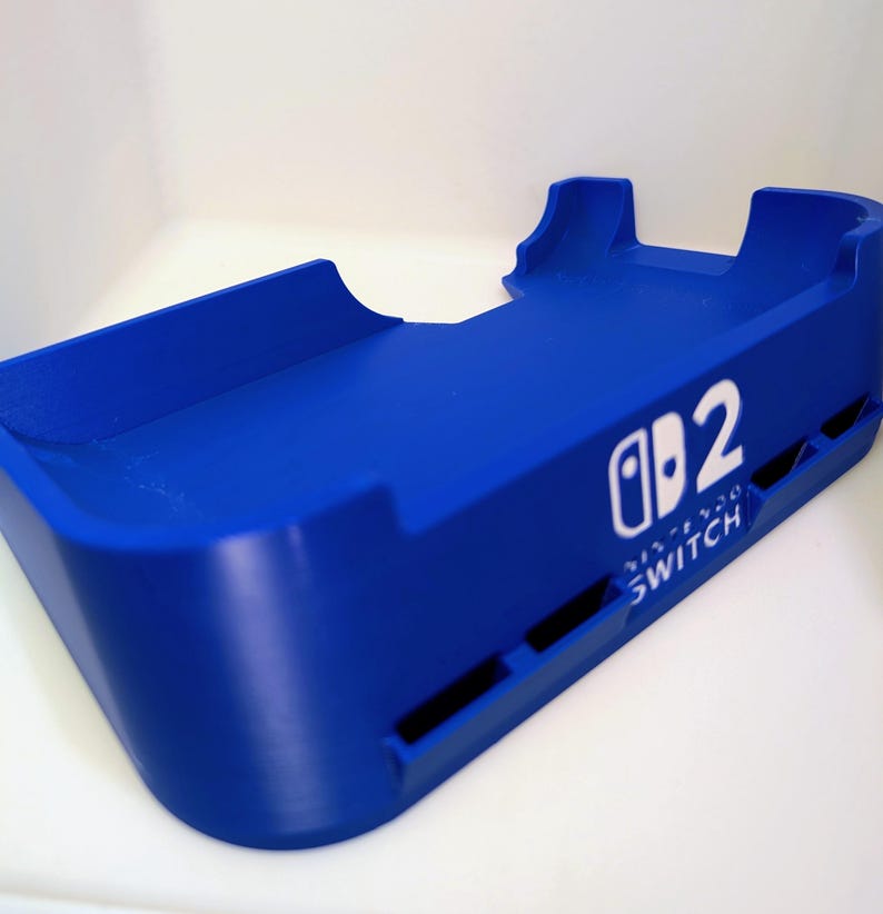 Może przedstawiać: Żywy niebieski dock Nintendo Switch. Dock posiada logo Nintendo Switch i cyfrę 2 w kolorze białym. Projekt obejmuje tacę i dwa podwyższone podpory. Widoczne jest r&oacute;wnież słowo "SWITCH".