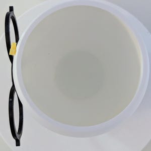 May include: A white ceramic cup with a black plastic rim and a yellow plastic handle. The cup is empty and the handle is on the left side of the image.