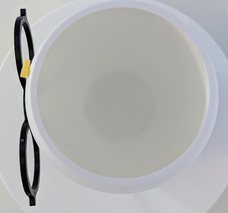 May include: A white ceramic cup with a black plastic rim and a yellow plastic handle. The cup is empty and the handle is on the left side of the image.