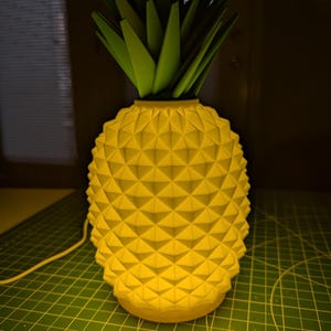 Puede incluir: Una lámpara amarilla con forma de piña y un patrón geométrico. La lámpara tiene una parte superior verde que se asemeja a hojas. La lámpara está enchufada e iluminada, emitiendo un brillo cálido. La lámpara está sobre una esterilla de corte verde.