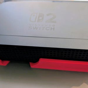 Może przedstawiać: Czarna konsola Nintendo Switch z dołączonymi szarymi kontrolerami. Konsola spoczywa na czerwonym stojaku. Tekst "Nintendo Switch" jest widoczny na konsoli.