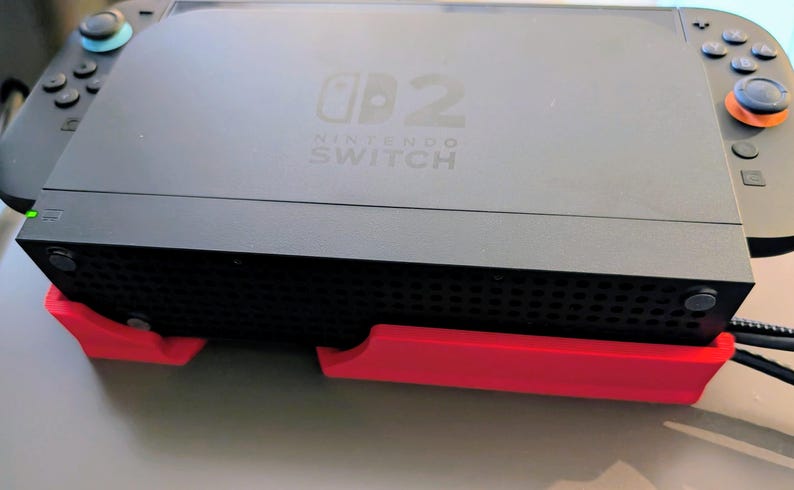 Może przedstawiać: Czarna konsola Nintendo Switch z dołączonymi szarymi kontrolerami. Konsola spoczywa na czerwonym stojaku. Tekst "Nintendo Switch" jest widoczny na konsoli.