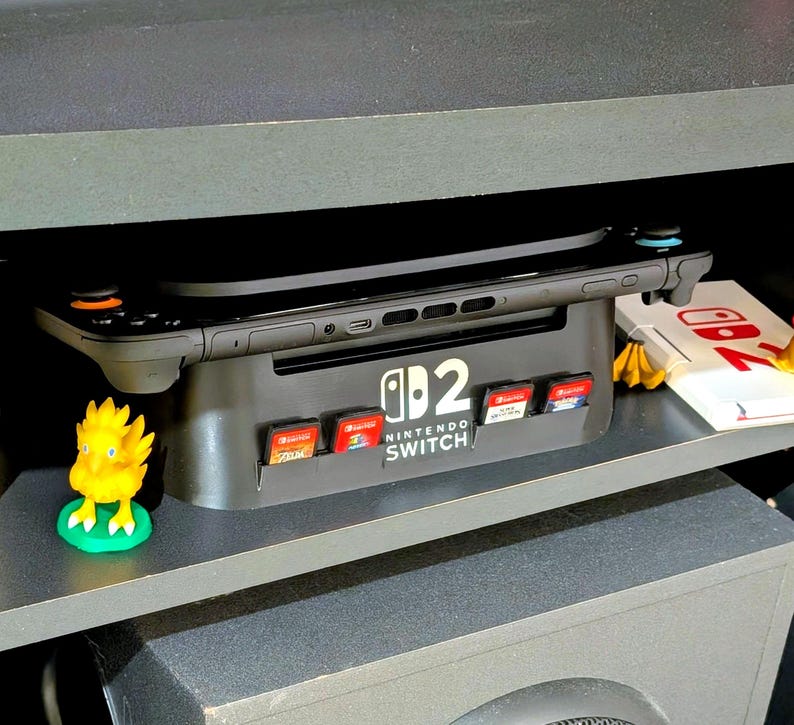 May include: A black Nintendo Switch console rests on a gray stand with game cartridge slots. The stand has the Nintendo Switch logo. Several game cartridges are inserted. A yellow Chocobo figurine sits on the shelf below. The console is on a shelf with a white box with red accents.