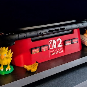 Może przedstawiać: Czerwona stacja dokująca Nintendo Switch z logo Nintendo Switch i kartridżami do gier. Czarna konsola Nintendo Switch jest zadokowana. Ż&oacute;łta figurka Chocobo i banan znajdują się na pierwszym planie.