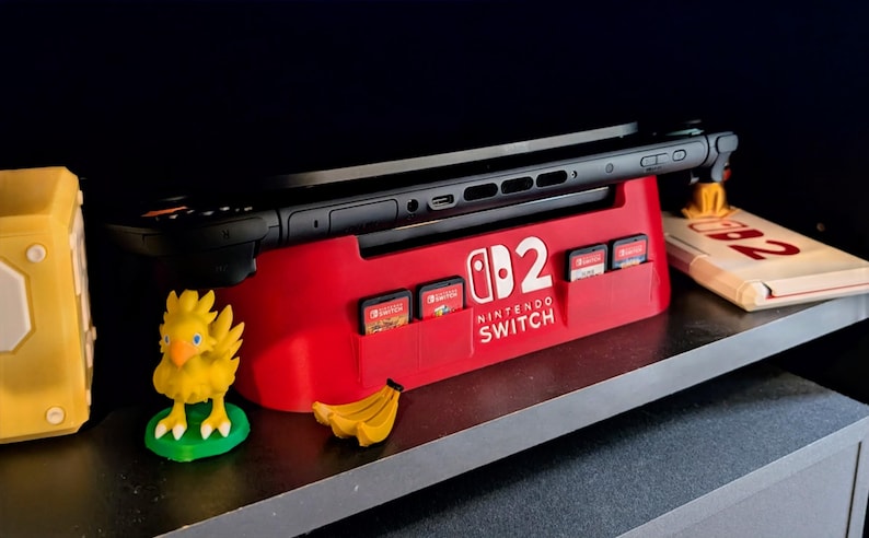 Może przedstawiać: Czerwona stacja dokująca Nintendo Switch z logo Nintendo Switch i kartridżami do gier. Czarna konsola Nintendo Switch jest zadokowana. Ż&oacute;łta figurka Chocobo i banan znajdują się na pierwszym planie.