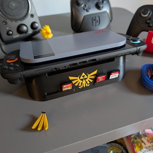 Może przedstawiać: Czarna stacja dokująca do Nintendo Switch z złotym herbem Zeldy i gniazdami na kartridże do gier. Szara konsola znajduje się na g&oacute;rze, z czarnym kontrolerem po lewej i czarno-czerwonym po prawej. Mała ż&oacute;łta figurka banana na pierwszym planie.