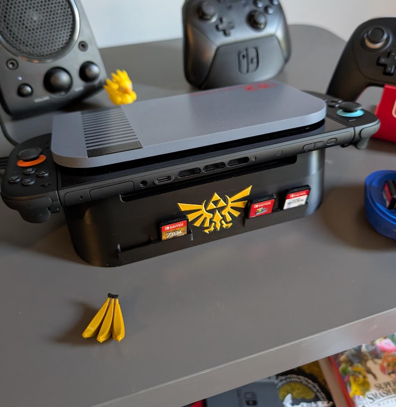 Może przedstawiać: Czarna stacja dokująca do Nintendo Switch z złotym herbem Zeldy i gniazdami na kartridże do gier. Szara konsola znajduje się na g&oacute;rze, z czarnym kontrolerem po lewej i czarno-czerwonym po prawej. Mała ż&oacute;łta figurka banana na pierwszym planie.