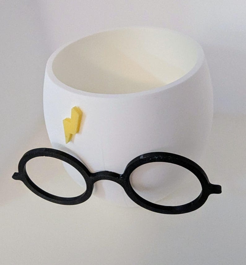 May include: White ceramic planter shaped like a head with a yellow lightning bolt and black round glasses.