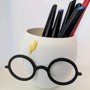 May include: White ceramic pencil holder shaped like a face with round black glasses and a yellow lightning bolt sticker. The holder is filled with black and red pens.