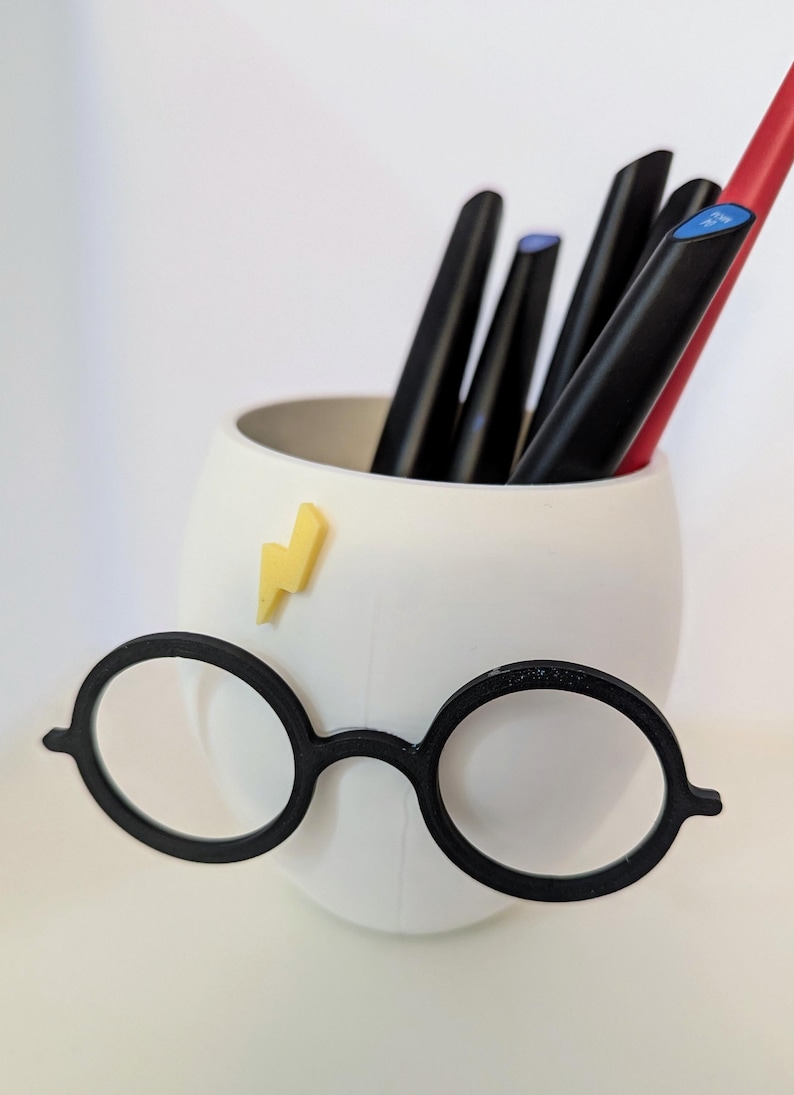 May include: White ceramic pencil holder shaped like a face with round black glasses and a yellow lightning bolt sticker. The holder is filled with black and red pens.