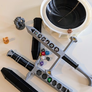 Puede incluir: Un conjunto de portadados de plástico gris con forma de espadas, una torre de dados de plástico negro y una bandeja de dados de plástico blanco con un centro negro. Hay varios dados de colores esparcidos alrededor de los portadados.