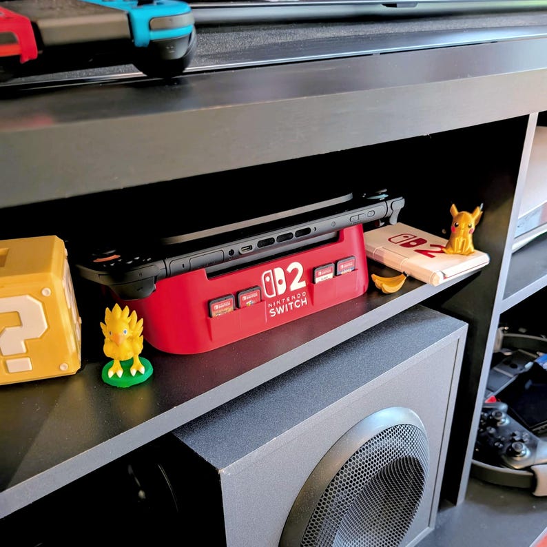 Może przedstawiać: Czerwona stacja dokująca Nintendo Switch z napisem "Nintendo Switch". Mieści czarną konsolę Nintendo Switch. Widoczny jest r&oacute;wnież ż&oacute;łty blok pytajnika Super Mario i figurka Pikachu.