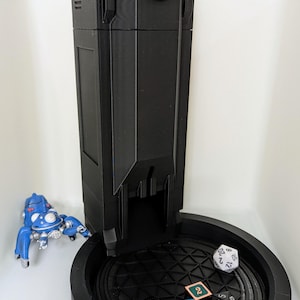 May include: A black 3D printed dice tower with a white symbol on top. The tower is sitting on a black circular tray with a geometric pattern. There are several dice on the tray, including a black d6, a blue d8, a green d4, a white d20, and a green d6.
