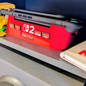 Może przedstawiać: Czerwona stacja dokująca Nintendo Switch z czarną konsolą Switch i kartridżami do gier. Widoczny jest r&oacute;wnież ż&oacute;łty blok Super Mario, ż&oacute;łta figurka ptaka i figurka Pikachu. Na p&oacute;łce znajduje się biała książka z logo Nintendo Switch.