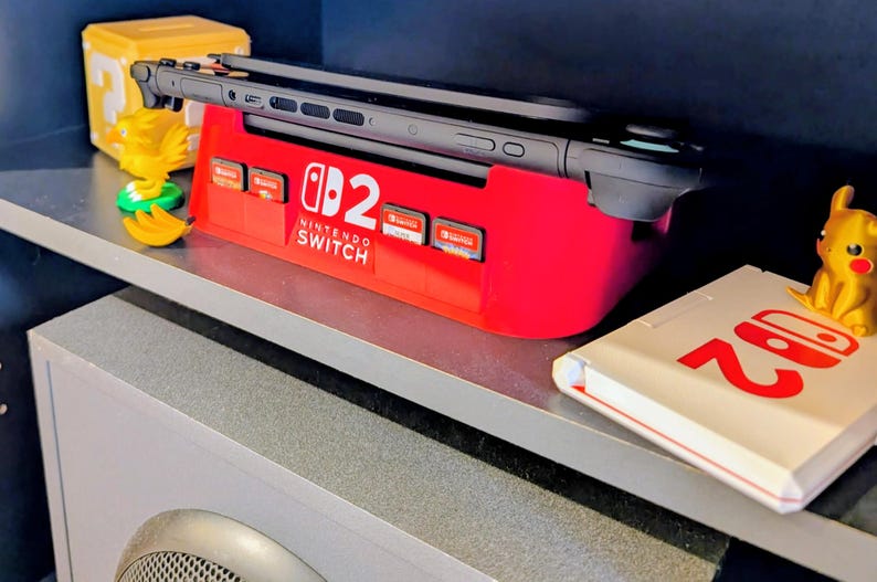 Może przedstawiać: Czerwona stacja dokująca Nintendo Switch z czarną konsolą Switch i kartridżami do gier. Widoczny jest r&oacute;wnież ż&oacute;łty blok Super Mario, ż&oacute;łta figurka ptaka i figurka Pikachu. Na p&oacute;łce znajduje się biała książka z logo Nintendo Switch.