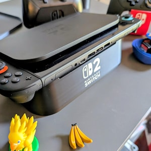 Może przedstawiać: Czarna konsola Nintendo Switch z dołączonym kontrolerem, spoczywająca na czarnej stacji ładującej. Na stacji dokującej widnieje napis "Nintendo Switch". Mała ż&oacute;łta figurka i pęk banan&oacute;w znajdują się na pierwszym planie.