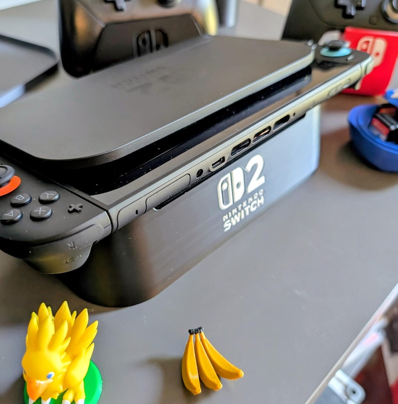 Może przedstawiać: Czarna konsola Nintendo Switch z dołączonym kontrolerem, spoczywająca na czarnej stacji ładującej. Na stacji dokującej widnieje napis "Nintendo Switch". Mała ż&oacute;łta figurka i pęk banan&oacute;w znajdują się na pierwszym planie.