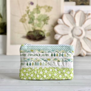 May include: A small, rectangular, quilted pouch with a white zipper. The pouch features various fabric patterns in shades of green and white, including floral, geometric, and teardrop designs. The text "Cotton Pattern # 30395" is printed on the fabric.