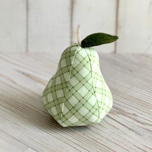 May include: A pear-shaped fabric ornament featuring a light green and white plaid pattern. The object has a dark green felt leaf attached to the top and a small stem protruding from the center, constructed with visible vertical seams.